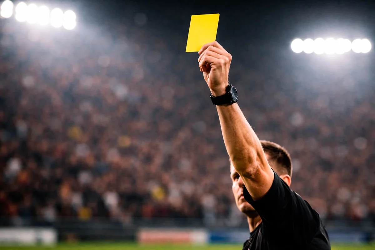Árbitro de fútbol mostrando una tarjeta amarilla en un campo de césped bajo los focos